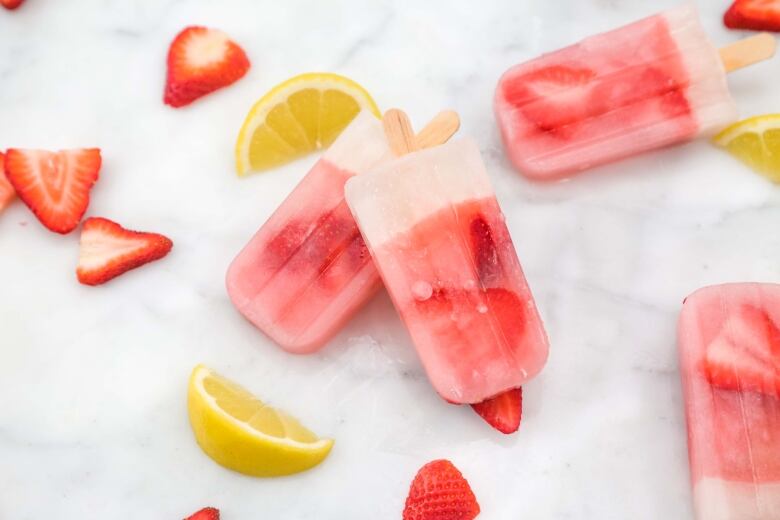 overhead shot of 4 pink popsicles with strawberry slices in them. strawberry and orange slices lay next to them on a white marble surface. 