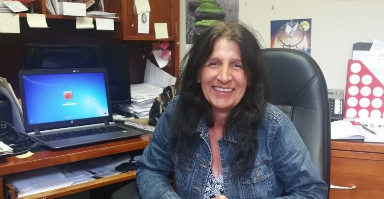 A woman with long black hair wearing a denim jacket sits at an office desk with a computer.