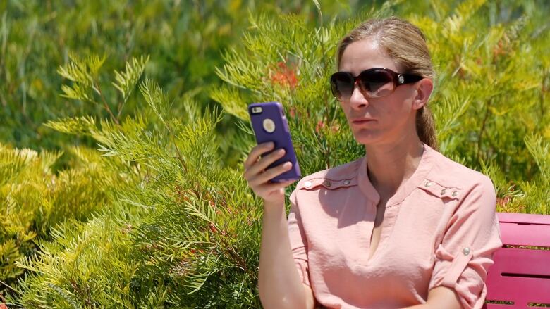 A woman in a salmon shirt and sunglasses looks at her phone.