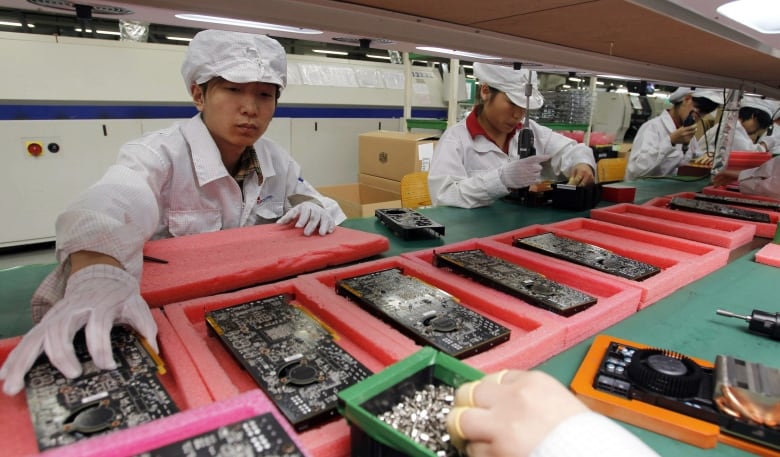 A uniformed worker wearing gloves reaches for a part on an circuit board assembly line as others work around him. 