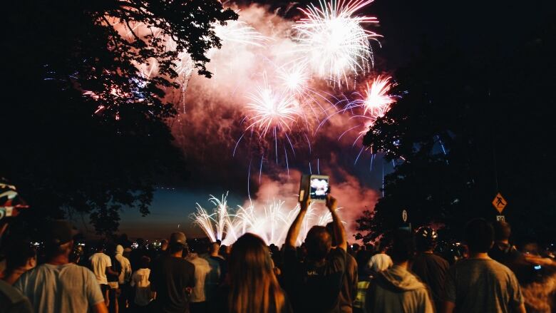 Fireworks light up the night sky, with hundreds of people visible in the foreground attempting to take pictures and videos.