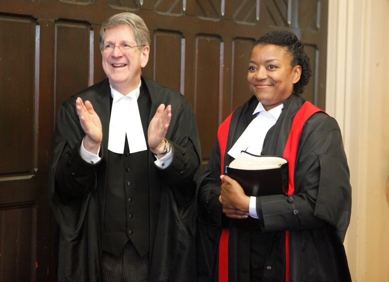 A male judge in black robes claps his hands for a female judge also in black robes standing to his right.