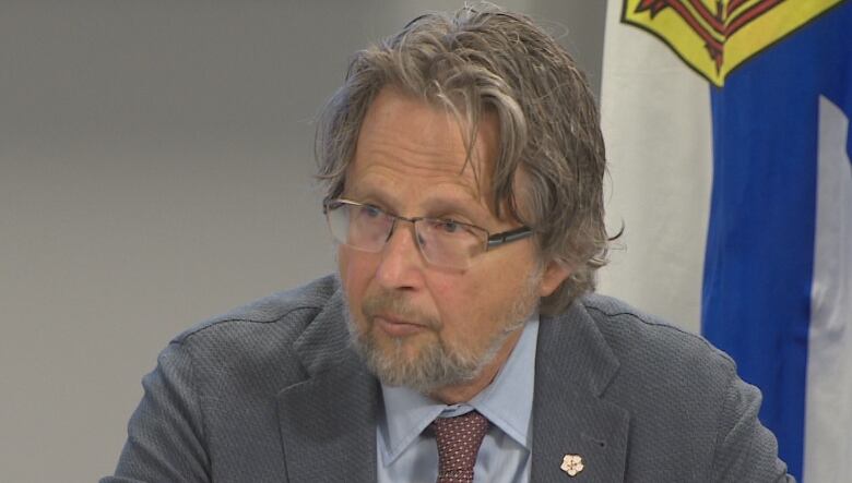 A man wearing glasses looks left and leans toward a small microphone. A Nova Scotia flag can be seen behind him.