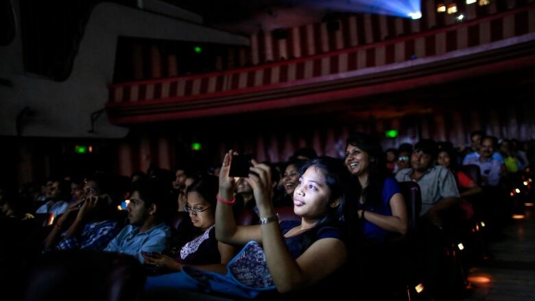 A girls face is illuminated by her phone screen as she takes a photo in a movie theatre. Behind her is a woman laughing while watching the screen. 