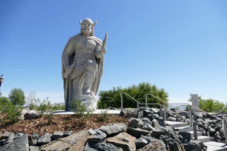 A statue of a 15 foot viking wearing a helmet and holding a staff towers over a small round park space against a blue sky. There are rocks and bushes surrounding him.  