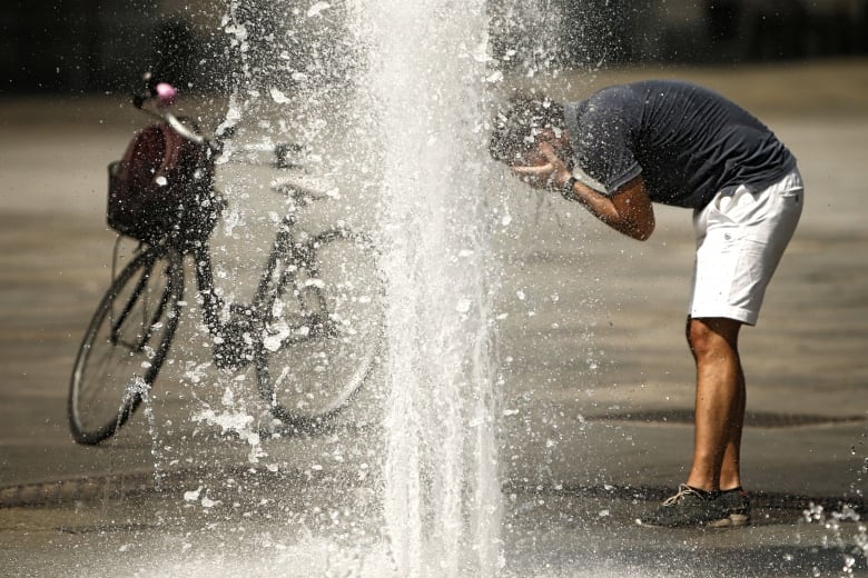 A man bends over with his hands on his face as he uses water from a fountain to cool down. His bicycle is nearby.