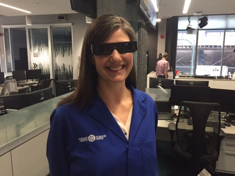 A woman wears a blue lab coat and dark glasses.