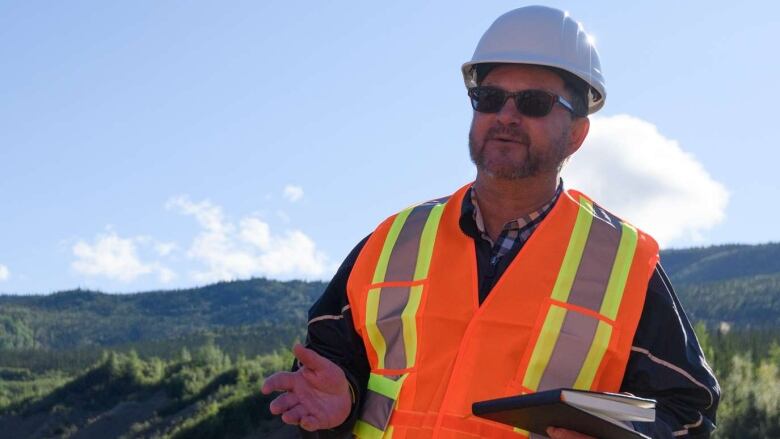 A man in a safety vest and hardhat stands outside in a remote, hilly area.