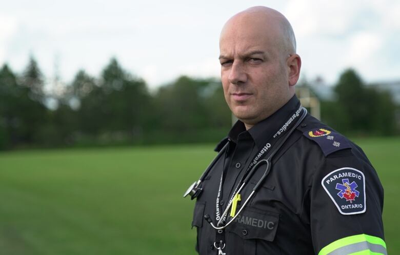 A bald man wearing a paramedic uniform, with a stethoscope around his neck, stands in a grassy field.