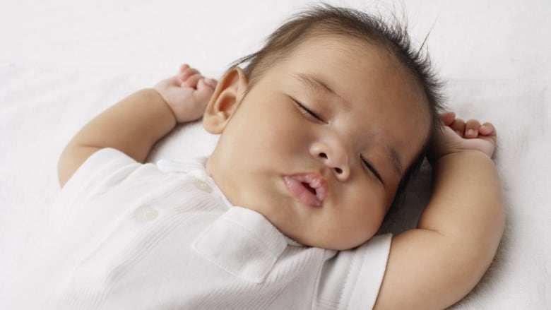 A closeup of a baby in white, fast asleep with their head tilted to their left and their arms raised to the sides of their head.