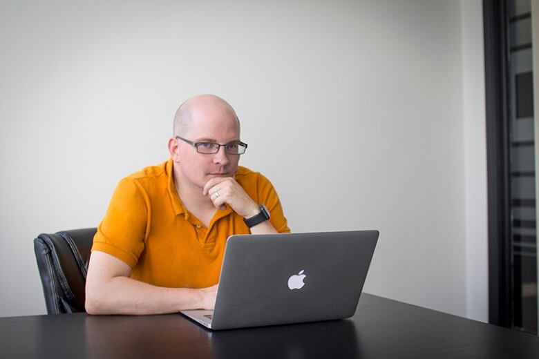 A bald man in an orange shirt on a laptop.