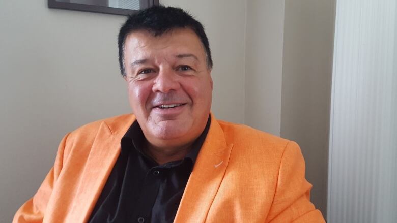 A man with dark hair, wearing a black shirt and orange jacket, sits and looks at the camera.