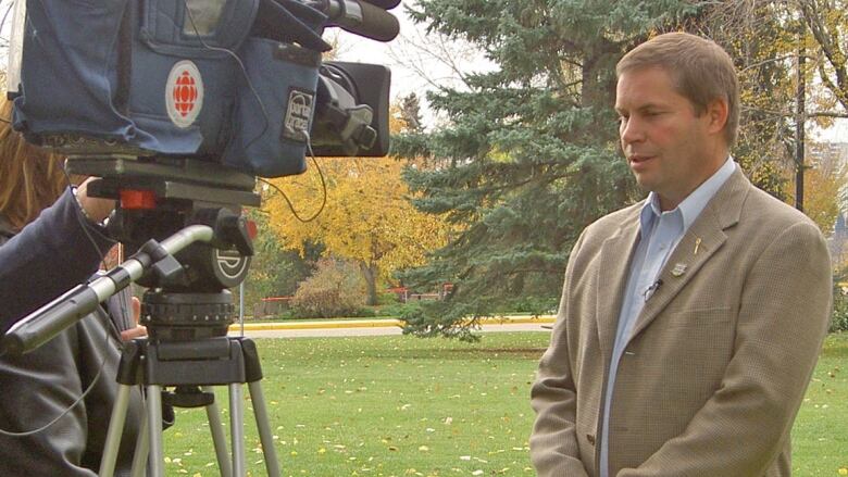 A white man in a tan suit with a blue shirt under it. He's standing outside and there is a television camera in front of him.