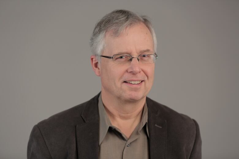 A photo shows a man with grey hair and glasses wearing a blazer and dress shirt.