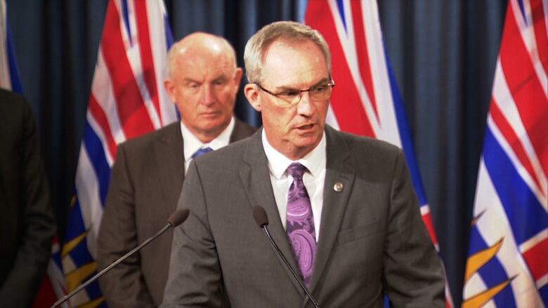 A white man speaks in front of a row of B.C. flags, with another white man behind him.