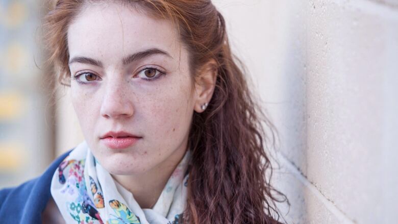 A young woman is photographed from the shoulders up. She wears a blue top and a white scarf with butterflies on it. She has a long red ponytail and is leaning against a white brick wall. 