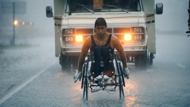 A man in vest rides with a bike for paraplegics in front of a vehicle amid rain.