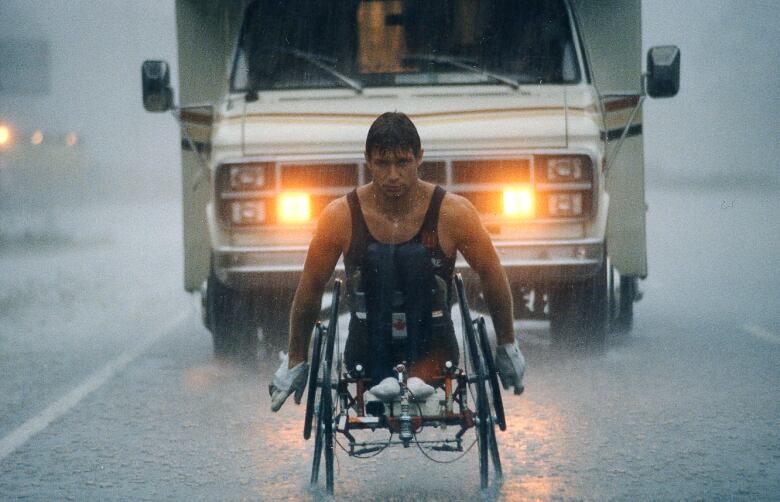 A man in vest rides with a bike for paraplegics in front of a vehicle amid rain.