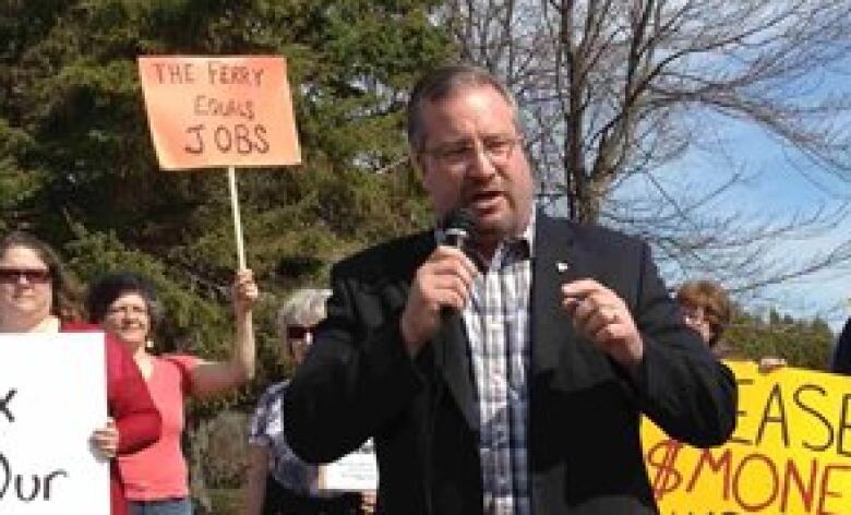 A man holding a microphone with protestors with signs in the background.