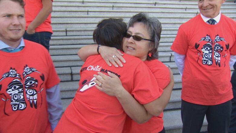 Two women, wearing orange shirts, hug.