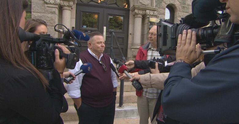 A person stands in front of a building, surrounded by people with cameras and microphones.