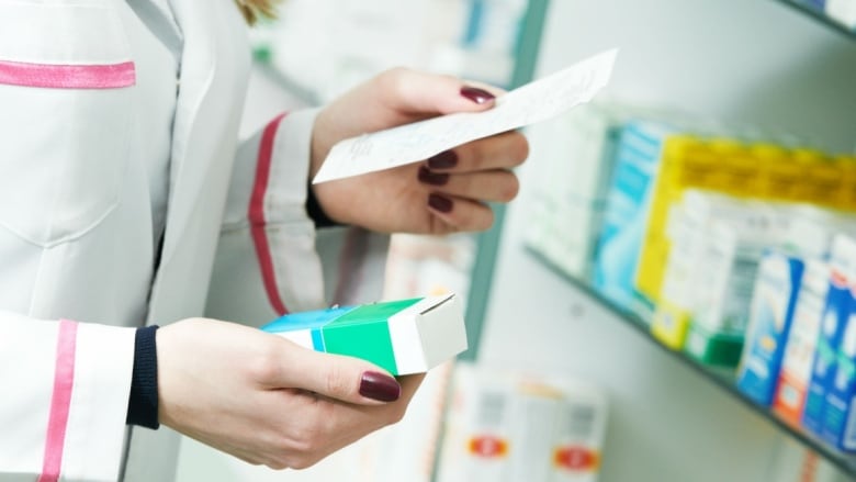 A closeup shows the hands of a woman wearing a white lab coat holding a box in front of a shelf lined with medication.