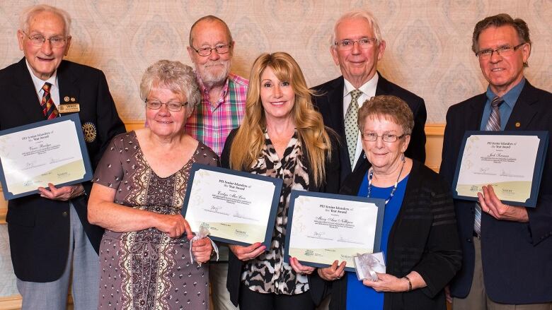 Seven people are shown posing for a photo, six of them holding framed certificates.