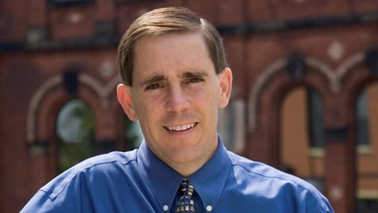 Man with brown hair in a blue shirt and tie stands in front of a brick building.