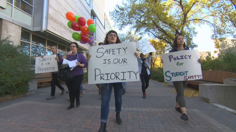 Students walk on a university campus carrying signs, with messages reading 