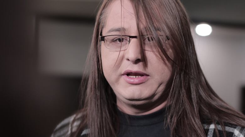 A woman with long brown hair and glasses looks toward the camera as she speaks.