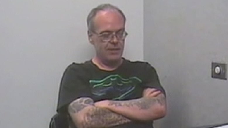 A man wearing a black t-shirt and glasses sits with his arms crossed on his chest. He has several tattoos on his arms.