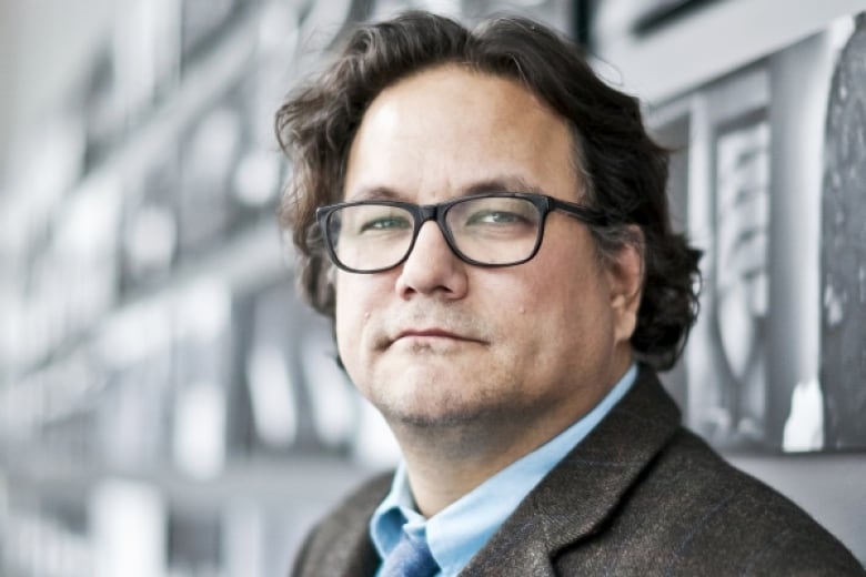 An close up of an Anishinaabe man wearing rectangular-framed glasses and his dark hair brushed back. He is wearing a suit and tie.