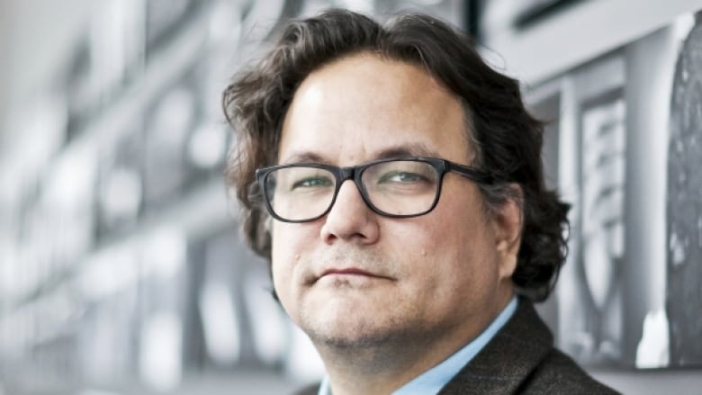 An close up of an Anishinaabe man wearing rectangular-framed glasses and his dark hair brushed back. He is wearing a suit and tie.