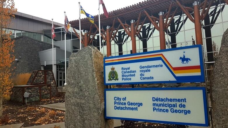 A sign announcing the City of Prince George RCMP detachment is seen next to a stone pillar outside a glass-walled building with trees showing their fall colours.