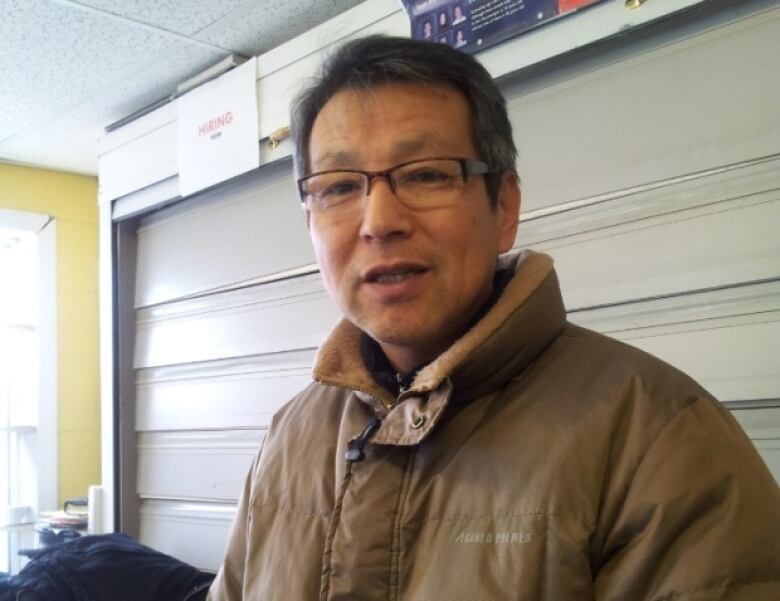 A man with dark hair and square glasses smiles at the camera. He is dressed in a light brown coat and appears to be behind the cash of a convenience store. 