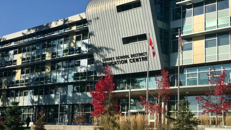 A grey building with a sign up front reads Surrey School District Education Centre. On the lawn in fron the of the building are red maple trees. 