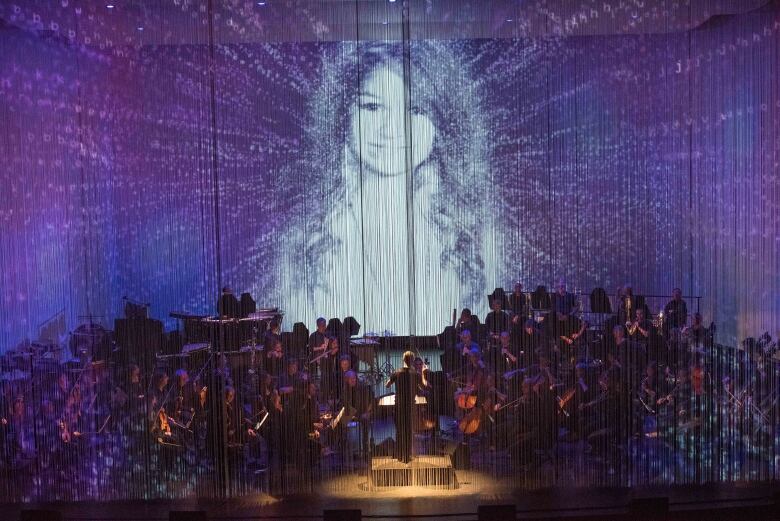 An orchestra performs while an ethereal image of Amanda Todd floats above the stage, surrounded by blue.