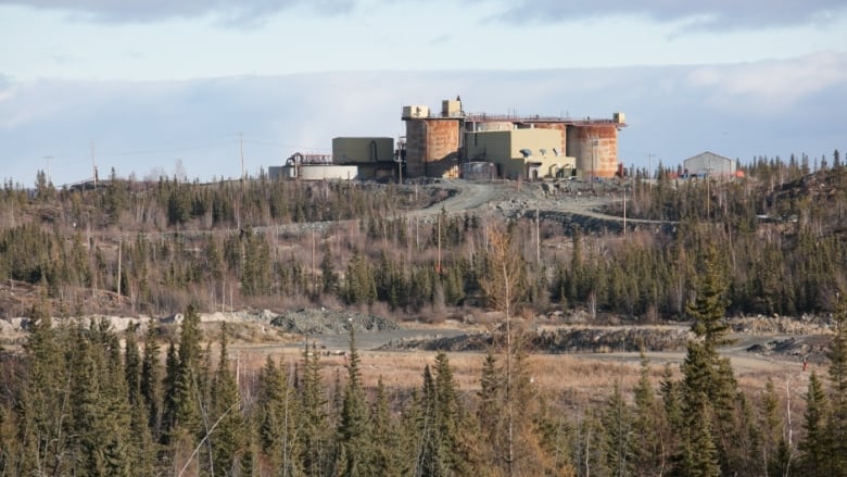 A building with huge round stack on the ends sits on top of a hill with trees and orange dirt in the foreground. 