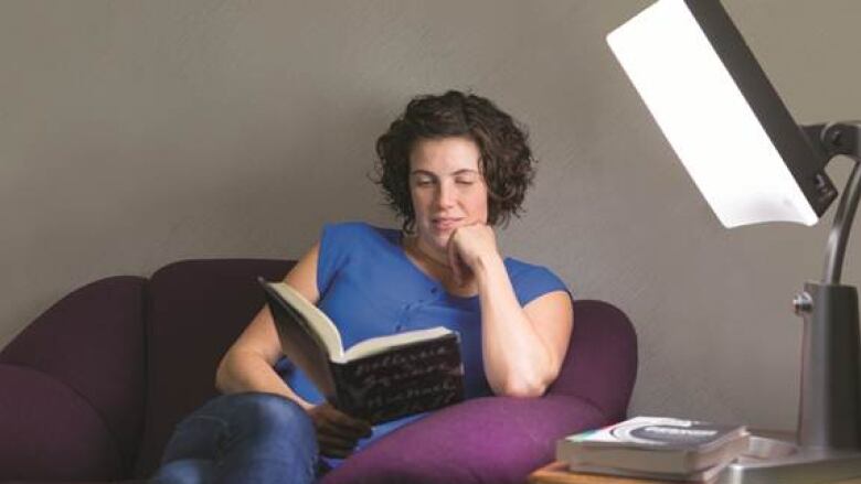 A woman sits in an armchair reading a book. A light therapy lamp is on next to her. 