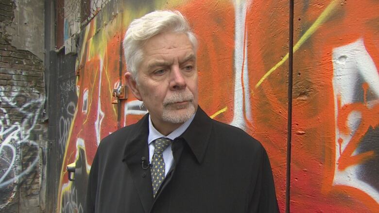 Headshot of Vancouver psychiatrist Dr. Bill MacEwan standing in front of an orange coloured wall with graffiti on it. 