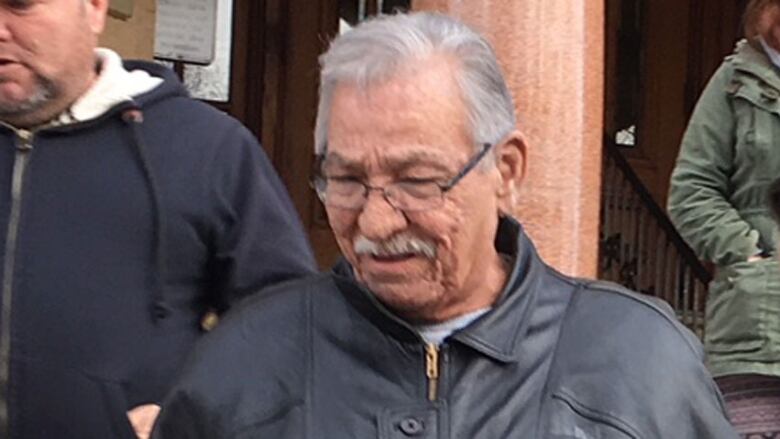 A portrait of a man with short grey hair and a moustache, wearing glasses and a black leather jacket, with his eyes downcast.