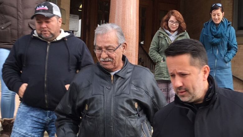 A man with short grey hair and a moustache, wearing glasses and a black leather jacket, with his eyes downcast, is flanked by two supporters, a man wearing a black coat, and another man wearing a hoodie, blue jeans and baseball cap.