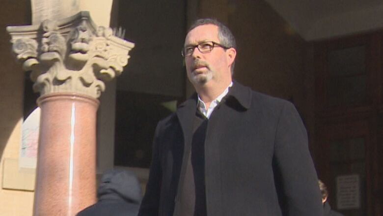 A low-angle portrait of a man with greying hair and a goatee, wearing glasses, a white collared shirt and black jacket, standing beside an ornate marble pillar.