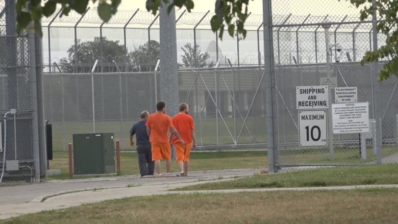 Two men in orange shorts and orange t-shirt walk beside a man in blue, in the shadow of large fences with barbed wire overtop. 