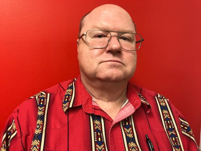 A picture of a man with glasses wearing a red shirt and there is a red background behind him. 