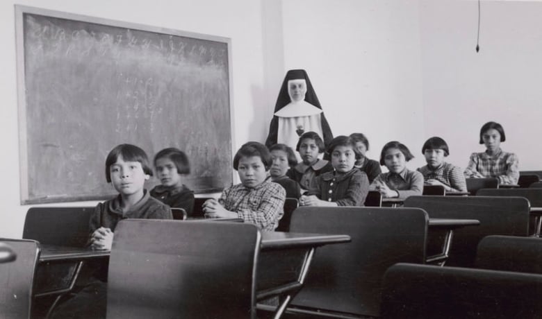 A black and white photo shows young students sitting in desks looking at the camera, with a woman in a nun's habit standing behind them.