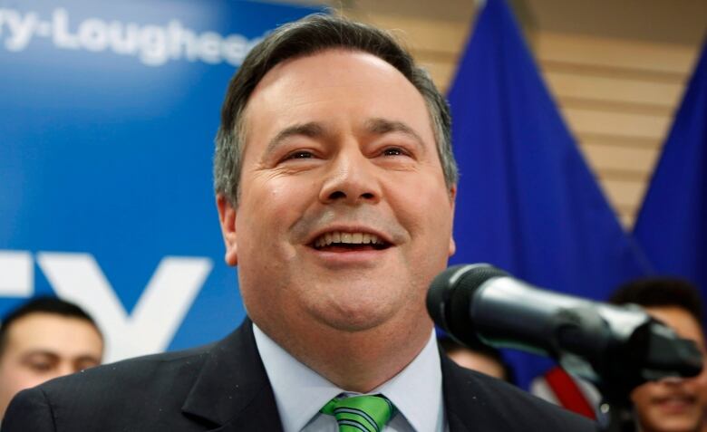 Jason Kenney smiles as he speaks into a microphone. He's wearing a dark suit and green tie.