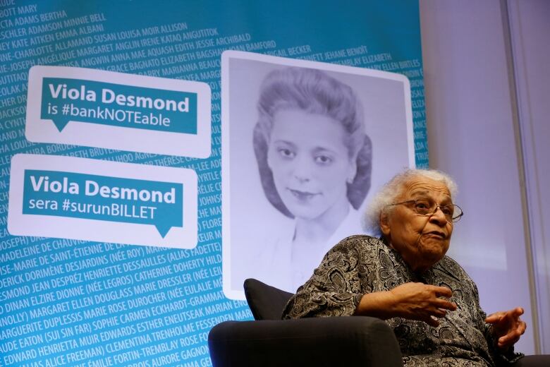 An older Black woman sits on a stage in front of an image of Viola Desmond