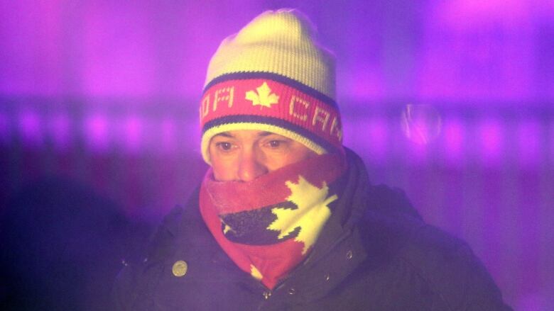 Someone in a tuque and neck warmer stands near a fire with a purple light glowing in the back.