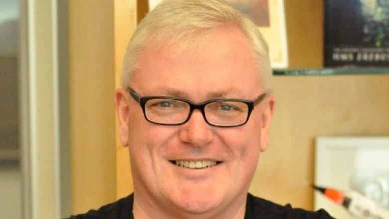 A man wearing black-rimmed glasses and a black T-shirt smiles as he poses for a photo.